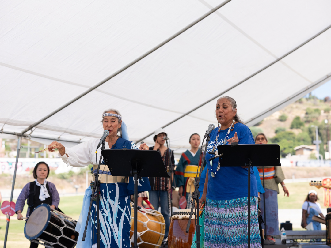 Two people stand on a stage within a white tent, each with a microphone in front of them. The person on the left wears a Kimono, featuring a white top and a blue, patterned skirt, with a headband. The person on the right wears a blue T-shirt and a multicolored, patterned skirt, and a shell necklace. Behind them, several people are present, dressed in traditional garments.